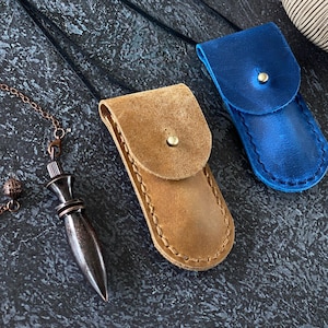 May include: Two leather pendant holders, one tan and one blue, with a button closure. A dark metal pendulum hangs from a chain on the left. The holders are on a dark, textured surface. A decorative object is in the background.