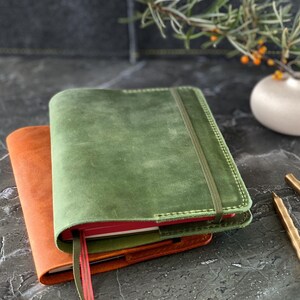 May include: Two leather-bound journals, one green and one orange, are stacked on top of each other. A gold pen is lying on a gray surface.