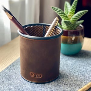 Puede incluir: Un portalápices de cuero marrón con una parte superior de malla, que contiene un lápiz y un bolígrafo. El portalápices tiene el nombre "Yaro" en relieve en la parte delantera. Una pequeña planta suculenta en maceta está cerca sobre una superficie gris.
