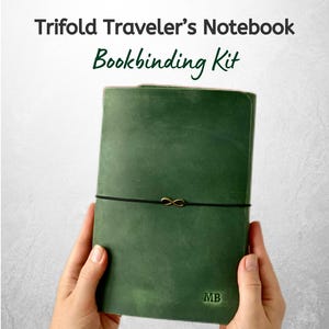 May include: A green trifold traveller's notebook bookbinding kit. The notebook is held closed with a black elastic cord and a gold-coloured infinity charm. The initials "MB" are embossed on the lower right corner. The text "Trifold Traveler's Notebook Bookbinding Kit" is at the top.