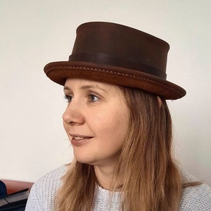 May include: A woman wearing a brown leather top hat with a stitched brim. She is wearing a light gray sweater.