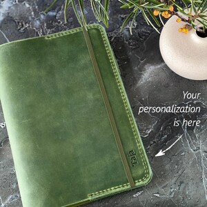 May include: A green leather journal with a stitched edge and a black elastic closure. The journal has the word "raja" embossed on the front cover. The text "Your personalization is here" is written on the image with an arrow pointing to the front cover. The text "Up 3 letters are FREE" is written below the journal.