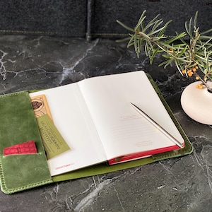May include: A green leather journal with a red pocket and a pen. The journal is open to a blank page. A small vase with a sprig of greenery is in the background.