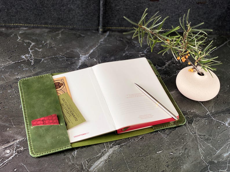 May include: A green leather journal with a red pocket and a pen. The journal is open to a blank page. A small vase with a sprig of greenery is in the background.