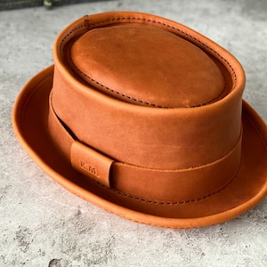 May include: A brown leather fedora hat with a stitched brim and a leather band with the initials "K.M." embossed on it.