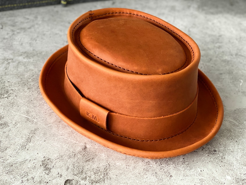 May include: A brown leather fedora hat with a stitched brim and a leather band with the initials "K.M." embossed on it.