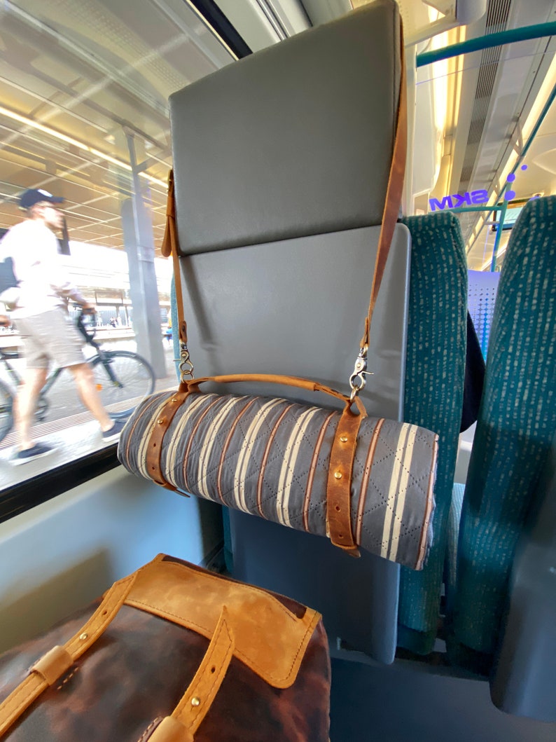 May include: A gray and white striped quilted blanket with brown leather straps is hanging from the back of a train seat. The blanket is rolled up and secured with the straps. A brown leather bag is sitting on the seat below the blanket.