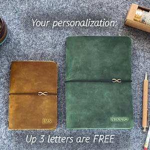 May include: Two leather journals, one brown and one green, with gold infinity symbol accents. The brown journal has the initials "JMS" and the green journal has the name "NIKKI" with a heart symbol. The text "Your personalization" and "Up 3 letters are FREE" are also visible.