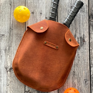 Puede incluir: Una bolsa de palas de cuero marr&oacute;n con dos palas negras en su interior. La bolsa tiene una solapa con cierre de presi&oacute;n y un peque&ntilde;o bolsillo en la parte delantera. La bolsa est&aacute; personalizada con el nombre "Michael".
