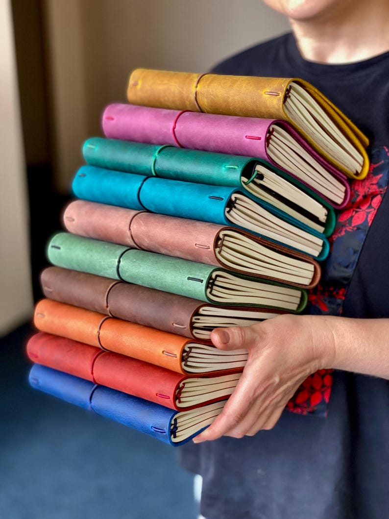 May include: A stack of colorful leather-bound journals in various shades, including yellow, pink, teal, blue, and brown. Each journal has a contrasting interior and a stitched spine. The journals are held in a person's hands.