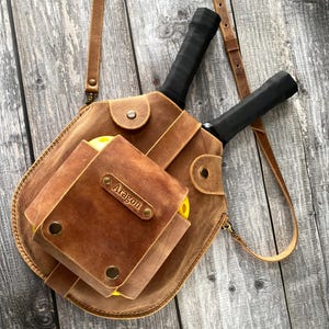 May include: A brown leather bag with a personalised name tag that says "Aragon". The bag has two pockets and a long strap. It is designed to hold two pickleball paddles and balls.