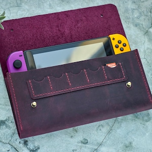Può includere: Una custodia in pelle bordeaux con una console Nintendo Switch all'interno. La custodia ha più slot per schede e una chiusura a lembo.