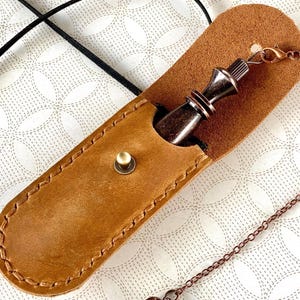 May include: A brown leather pendant case with a bronze chess king pendant. The case is stitched and has a press stud closure. A bronze chain is attached to the pendant. The background is a white pattern.