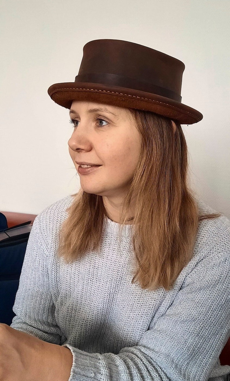 May include: A woman wearing a brown leather top hat with a stitched brim. She is wearing a light gray sweater.