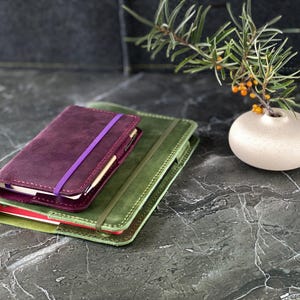 May include: Two leather-bound journals, one green and one burgundy, stacked on top of each other. The burgundy journal has a purple elastic band closure.