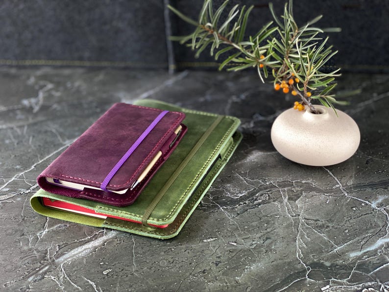 May include: Two leather-bound journals, one green and one burgundy, stacked on top of each other. The burgundy journal has a purple elastic band closure.