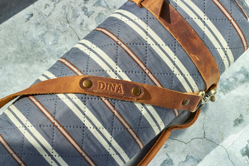 May include: A rolled up gray and white striped picnic blanket with a brown leather strap and buckle. The strap has a brass buckle and the name "DINA" is embossed on the leather.