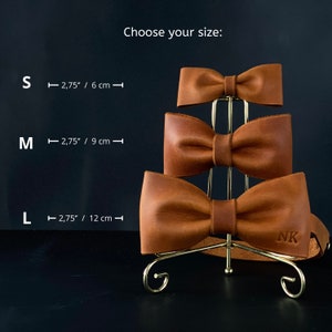 Può includere: Tre papillon in pelle marrone esposti su un supporto dorato. I papillon sono etichettati con le taglie S, M e L. Le misure per ogni taglia sono indicate in pollici e centimetri. Il papillon più grande ha le iniziali "NK" in rilievo.