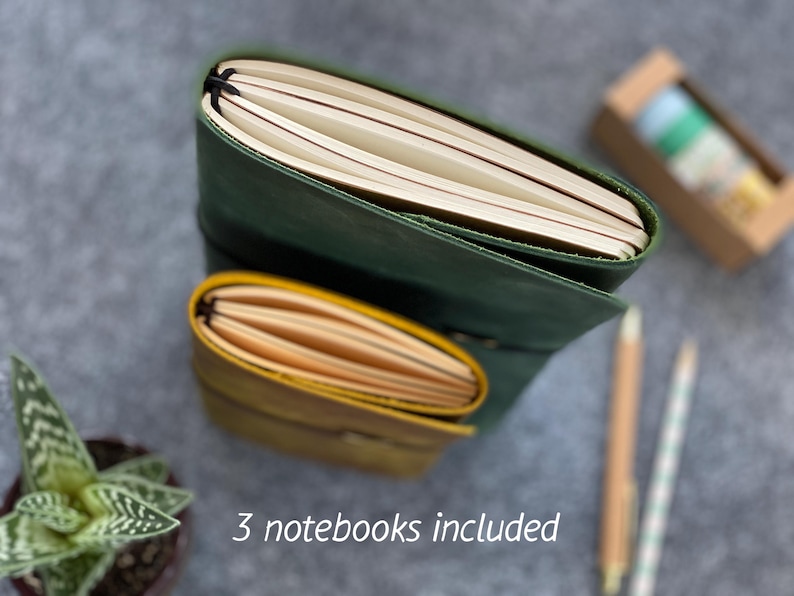 May include: Two leather notebooks, one green and one yellow, with cream-colored pages. The notebooks are stacked, with the green one on top. The text "3 notebooks included" is at the bottom. A small succulent plant, pencils, and washi tape are also visible.