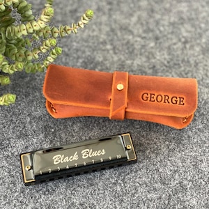 May include: A black harmonica with the text "Black Blues" and a brown leather case with the name "GEORGE" embossed on it. The harmonica has gold-colored accents and a row of numbered holes. The case is closed with a button.