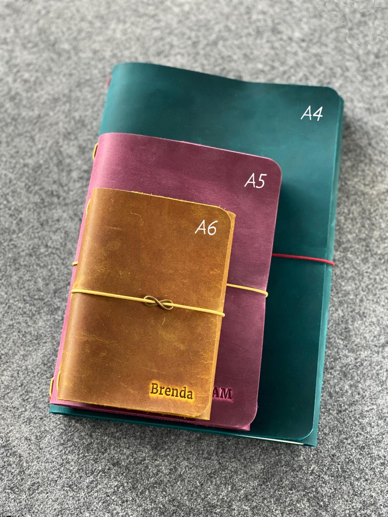 May include: Three leather journals in A4, A5, and A6 sizes. The A4 journal is teal, the A5 is burgundy, and the A6 is brown with the name "Brenda" embossed in gold. Each journal is bound with an elastic cord.