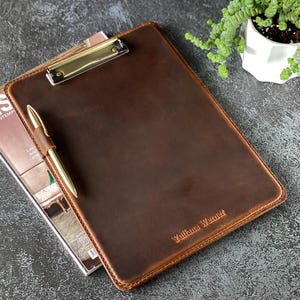 May include: A brown leather clipboard with a gold metal clip and a pen. The clipboard has the name "Yuliana Werner" embossed on the front.