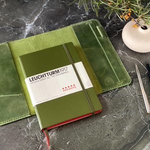 May include: A green leather notebook cover with a green notebook inside. The notebook has the text "LEUCHTTURM1917" and "DETAILS MAKE ALL THE DIFFERENCE" on the front cover. The notebook also has a red band around the edge and the text "RED DOTS" on the front cover.