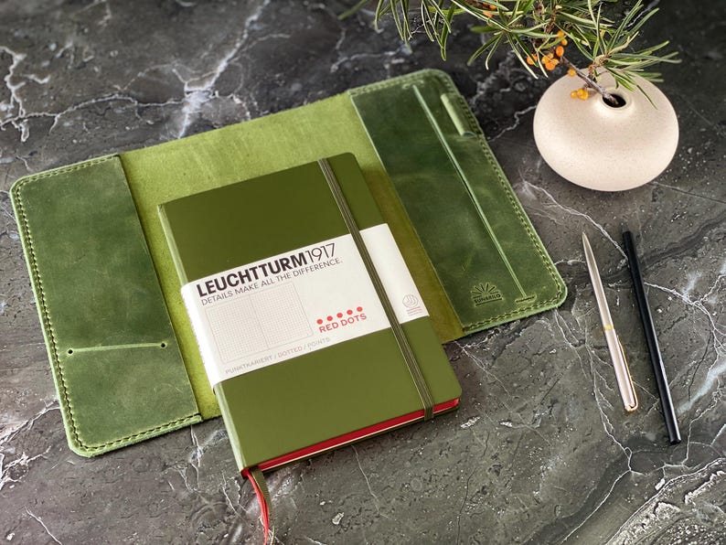 May include: A green leather notebook cover with a green notebook inside. The notebook has the text "LEUCHTTURM1917" and "DETAILS MAKE ALL THE DIFFERENCE" on the front cover. The notebook also has a red band around the edge and the text "RED DOTS" on the front cover.