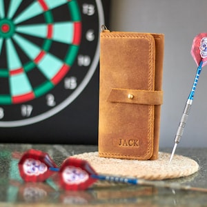 May include: A brown leather wallet with a flap closure and a gold button. The wallet has the name "JACK" embossed on the front. The wallet is sitting on a table with a dartboard and darts in the background.