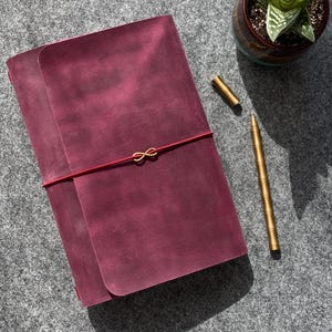 May include: A burgundy leather journal with a red elastic closure and gold-colored infinity charm. A gold pen and a small potted succulent are also visible. The journal is closed, and the scene is set on a gray surface.