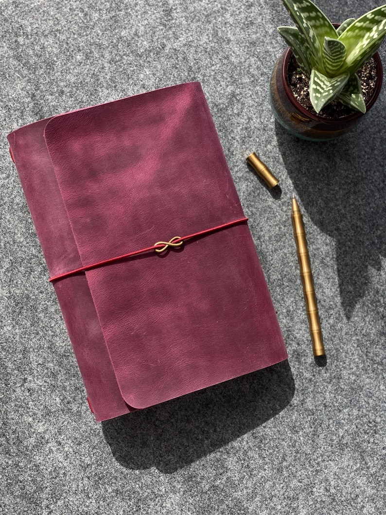 May include: A burgundy leather journal with a red elastic closure and gold-colored infinity charm. A gold pen and a small potted succulent are also visible. The journal is closed, and the scene is set on a gray surface.