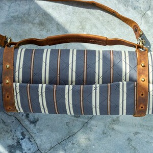 May include: A rolled-up striped picnic blanket with a brown leather strap and buckle. The blanket is made of a soft, quilted fabric and has a light blue and white striped pattern.