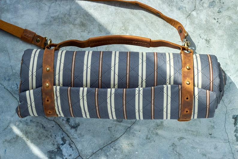 May include: A rolled-up striped picnic blanket with a brown leather strap and buckle. The blanket is made of a soft, quilted fabric and has a light blue and white striped pattern.