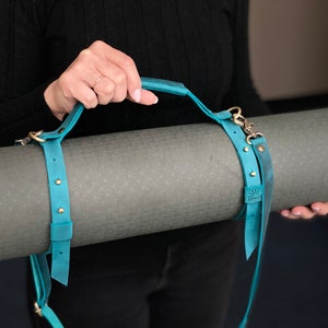 May include: A teal blue leather yoga mat strap with a handle and brass hardware. The strap is wrapped around a rolled up gray yoga mat.