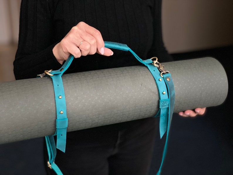 May include: A teal blue leather yoga mat strap with a handle and brass hardware. The strap is wrapped around a rolled up gray yoga mat.