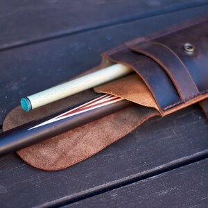 May include: A dark brown leather cue stick case with a snap closure, holding two pool cues. One cue is light-colored with a blue tip, the other is dark with red and white stripes. The case has a strap.