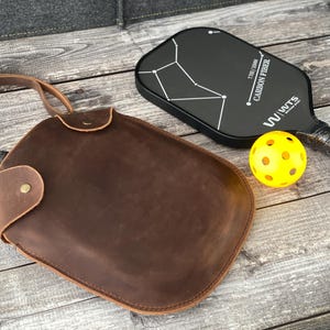 Puede incluir: Una bolsa de paleta de cuero marr&oacute;n con una correa para el hombro. La bolsa est&aacute; al lado de una paleta de pickleball negra con un dise&ntilde;o de constelaci&oacute;n blanca y el texto "TUBBY 1300mm CARBON FIBER W/WUTTS". Tambi&eacute;n hay una pelota de pickleball amarilla en la imagen.