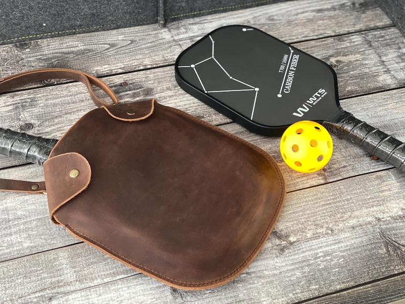Puede incluir: Una bolsa de paleta de cuero marr&oacute;n con una correa para el hombro. La bolsa est&aacute; al lado de una paleta de pickleball negra con un dise&ntilde;o de constelaci&oacute;n blanca y el texto "TUBBY 1300mm CARBON FIBER W/WUTTS". Tambi&eacute;n hay una pelota de pickleball amarilla en la imagen.