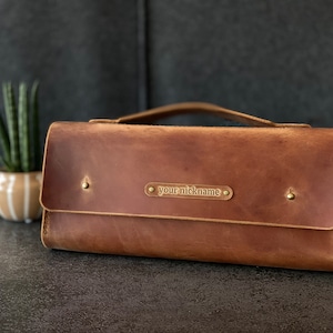 Könnte beinhalten: Braune Leder-Clutch mit Griff, Klappe und einem kleinen Metallschild mit der Aufschrift "your nickname". Die Tasche hat Messingnieten und ein Used-Finish. Eine kleine Pflanze in einem gestreiften Topf befindet sich im Hintergrund.