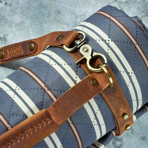 May include: A rolled up striped fabric picnic blanket with a brown leather strap and a brass clasp. The strap has the name "DINA" embossed on it.