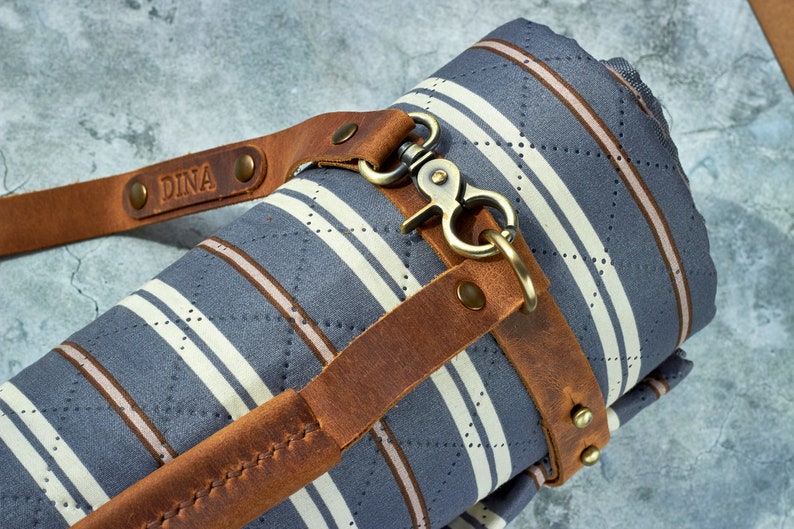 May include: A rolled up striped fabric picnic blanket with a brown leather strap and a brass clasp. The strap has the name "DINA" embossed on it.