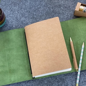 May include: A brown notebook with a kraft paper cover is open on a green leather surface. A gold pen and a striped pencil are next to the notebook. A small potted succulent and a box of decorative tape complete the scene.