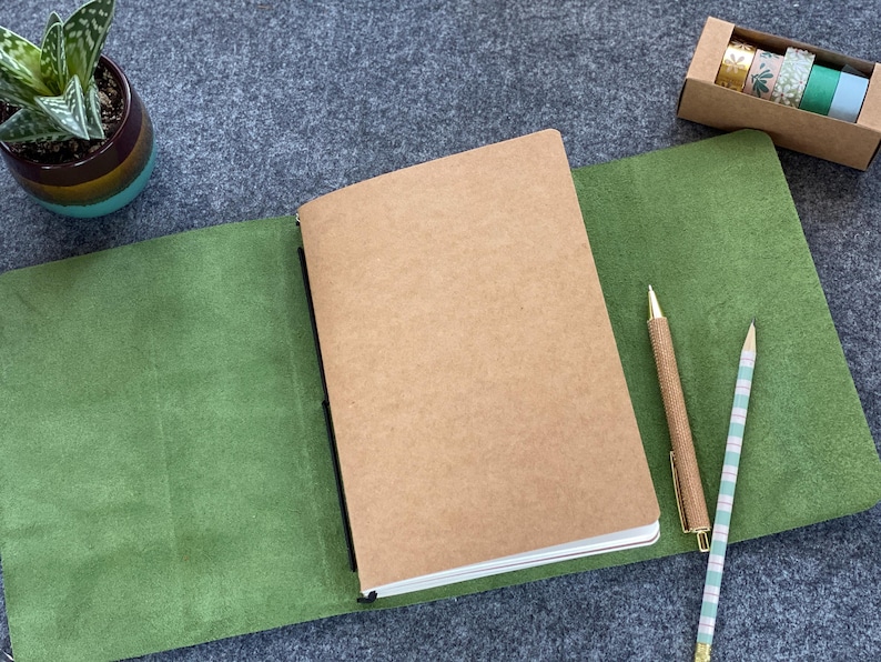 May include: A brown notebook with a kraft paper cover is open on a green leather surface. A gold pen and a striped pencil are next to the notebook. A small potted succulent and a box of decorative tape complete the scene.