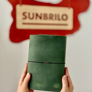 May include: A green leather journal with a black elastic closure and a gold infinity charm. The initials "MB" are embossed on the lower right corner. The word "SUNBRILO" is visible in the background.