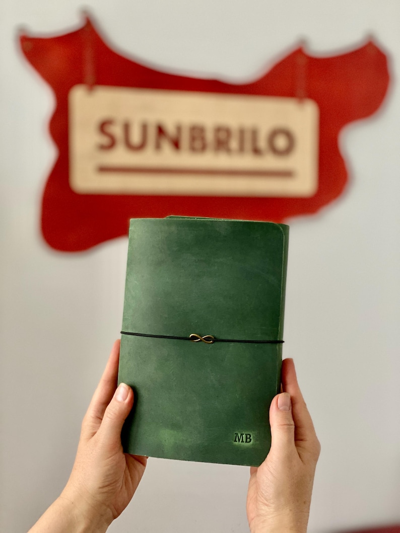 May include: A green leather journal with a black elastic closure and a gold infinity charm. The initials "MB" are embossed on the lower right corner. The word "SUNBRILO" is visible in the background.