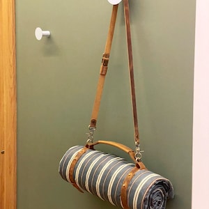 May include: A rolled up striped blanket with a brown leather strap and handle hanging from a white hook on a green wall.