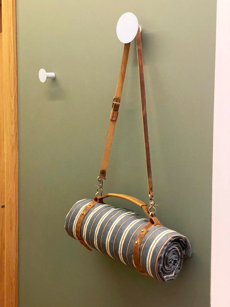 May include: A rolled up striped blanket with a brown leather strap and handle hanging from a white hook on a green wall.