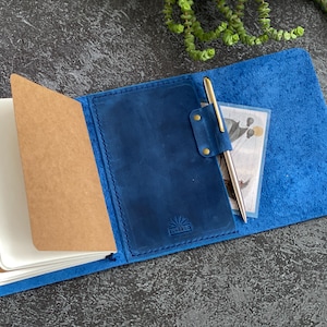 Op de afbeelding: Een blauwe leren notitieboekhoes met een ingebouwde penlus met een goudkleurige pen, een bruin papieren inzetstuk en een gestempeld logo. De binnenzijde bevat een vakje voor kleine voorwerpen, met zichtbare stiksels langs de randen van het leer.