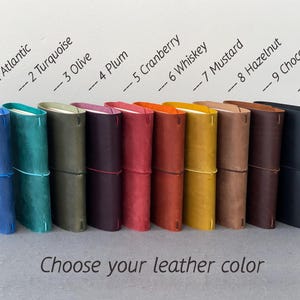 May include: A collection of ten leather journals in various colors, including blue, green, purple, red, orange, yellow, brown, and black. Each journal is secured with an elastic band. The text "Choose your leather color" is displayed below.