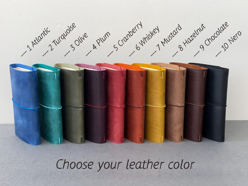 May include: A collection of ten leather journals in various colors, including blue, green, purple, red, orange, yellow, brown, and black. Each journal is secured with an elastic band. The text "Choose your leather color" is displayed below.